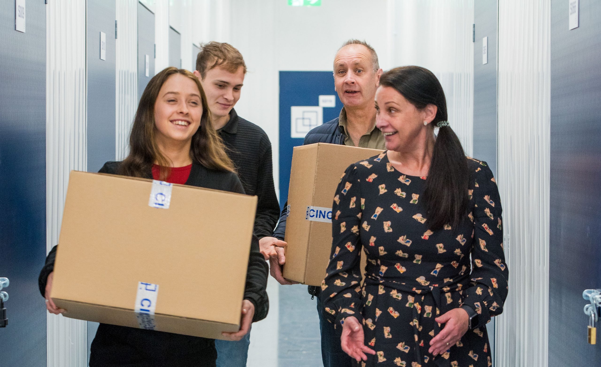 Two people carrying boxes while talking with their friends through a hallway