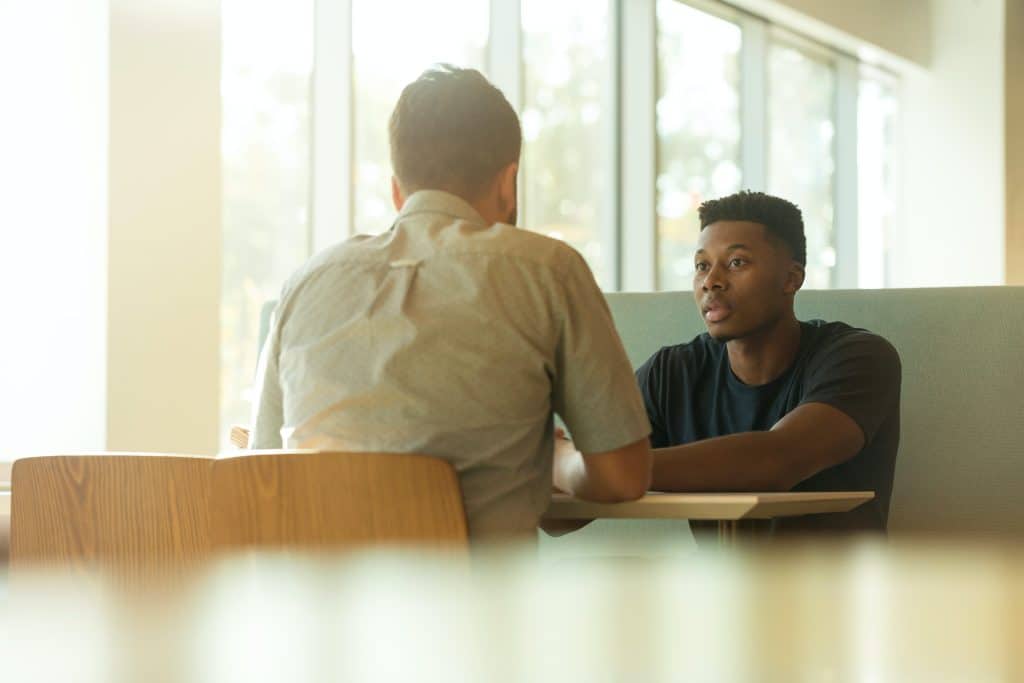 Two men conversing at a table by a window, warm light shining through