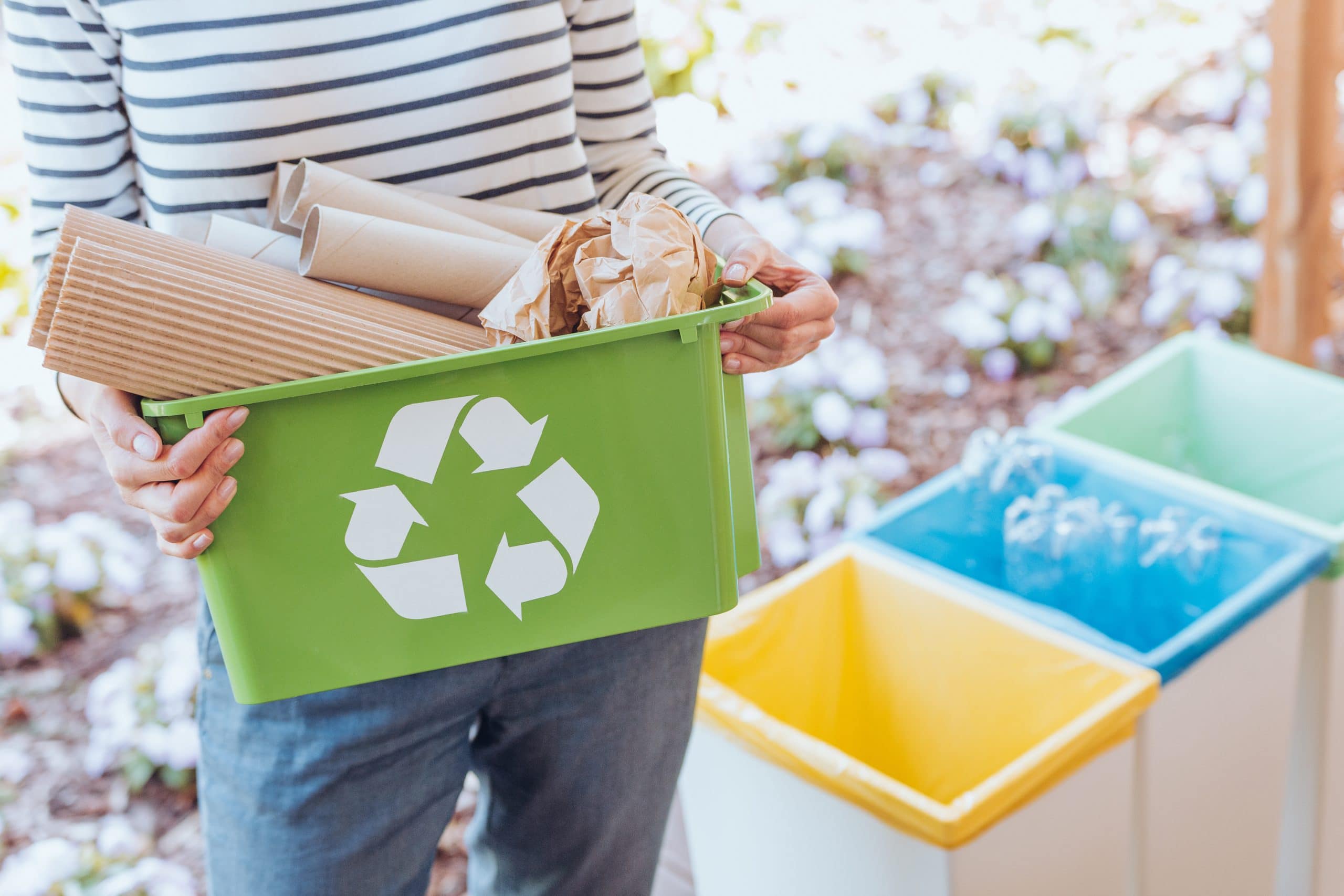 A person holding a green recycling box filled with cardboard. Other coloured bins next to her.