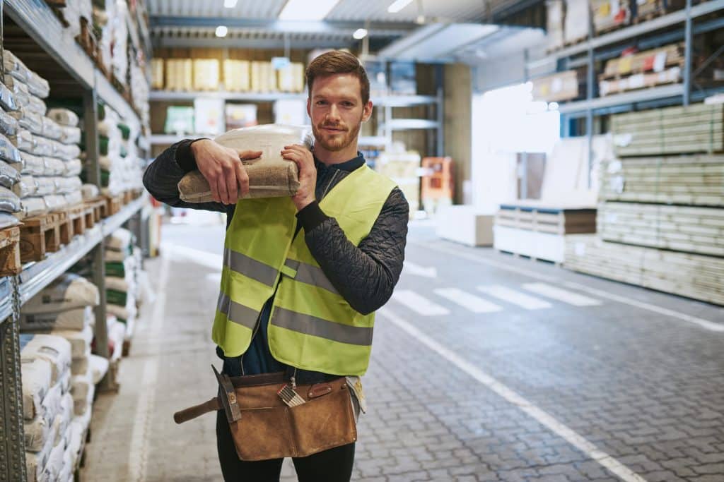 Builder in our storage for business unit surrounded by building materials and holding bag of cement on their shoulder