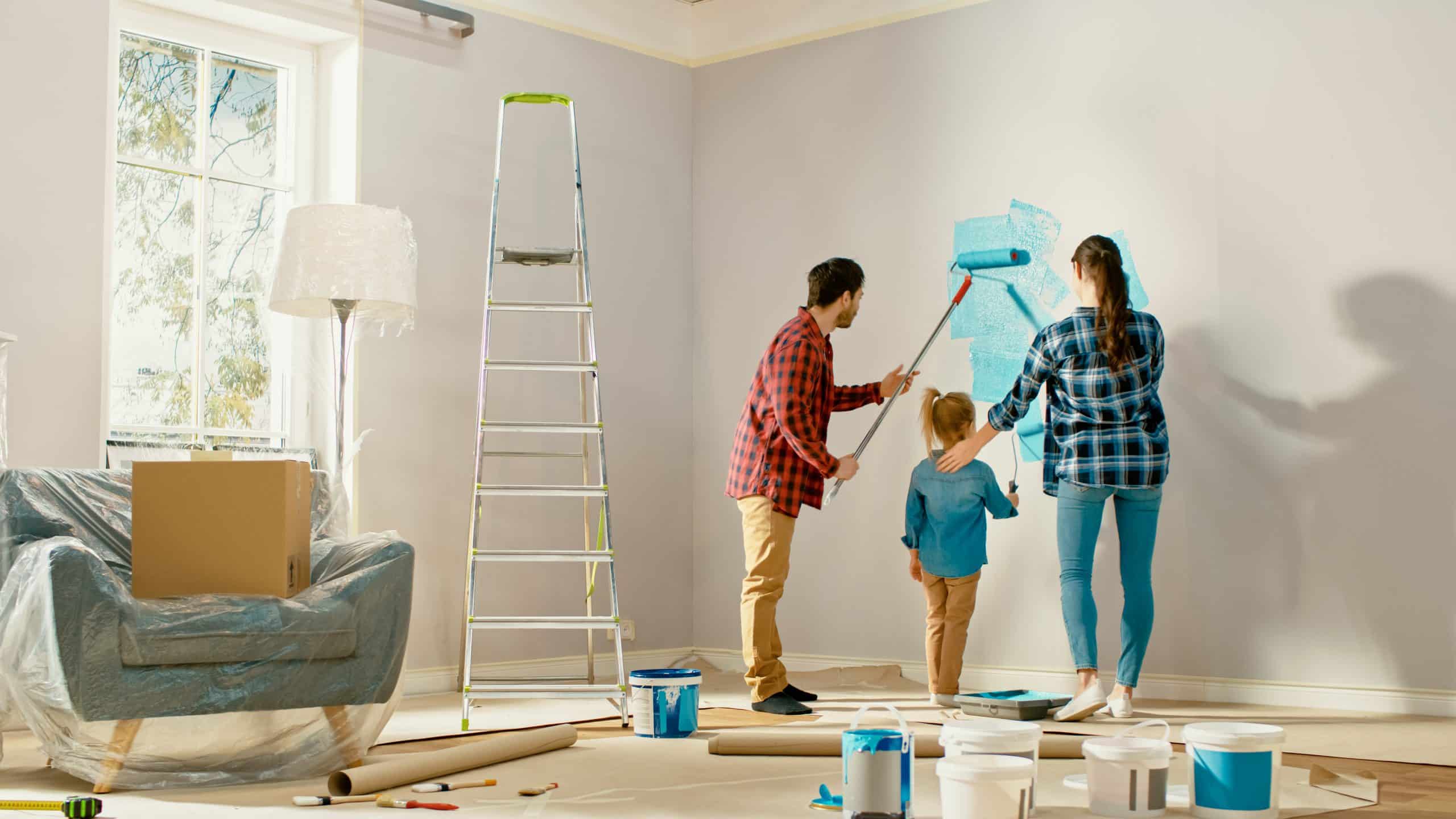 Shows the interior of a living room and a woman, man and child painting the wall in a blue coloured paint with furniture and boxes around them - self storage near Newbury can help during a home renovation