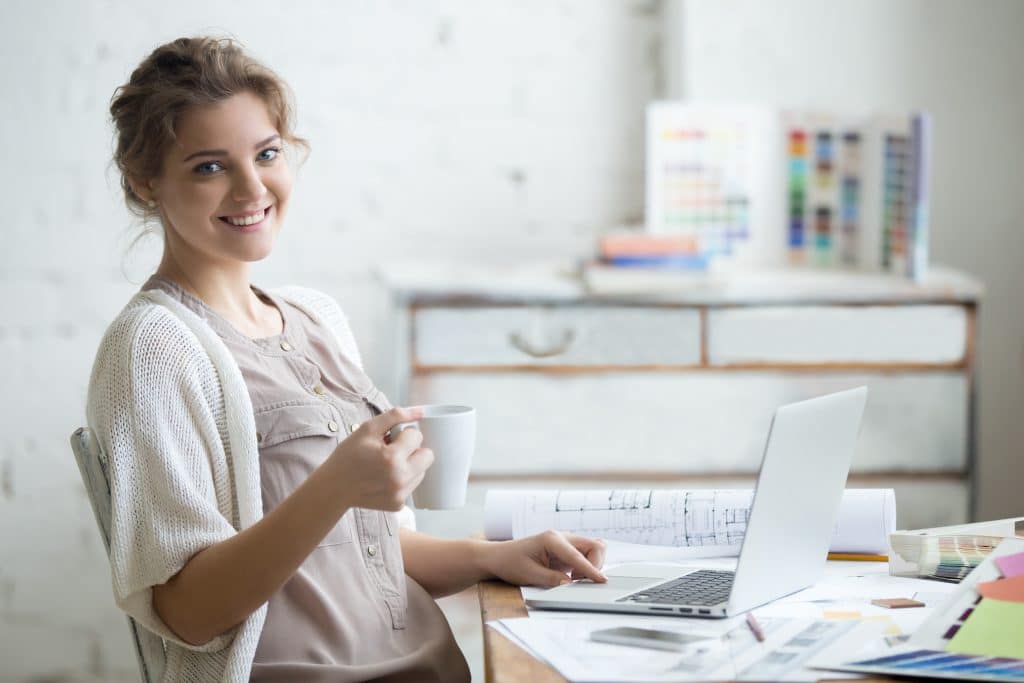 Office to rent Bicester - photo shows small business owner woman sitting at her desk, holding a cup of tea with house plans and an open laptop on the desk 