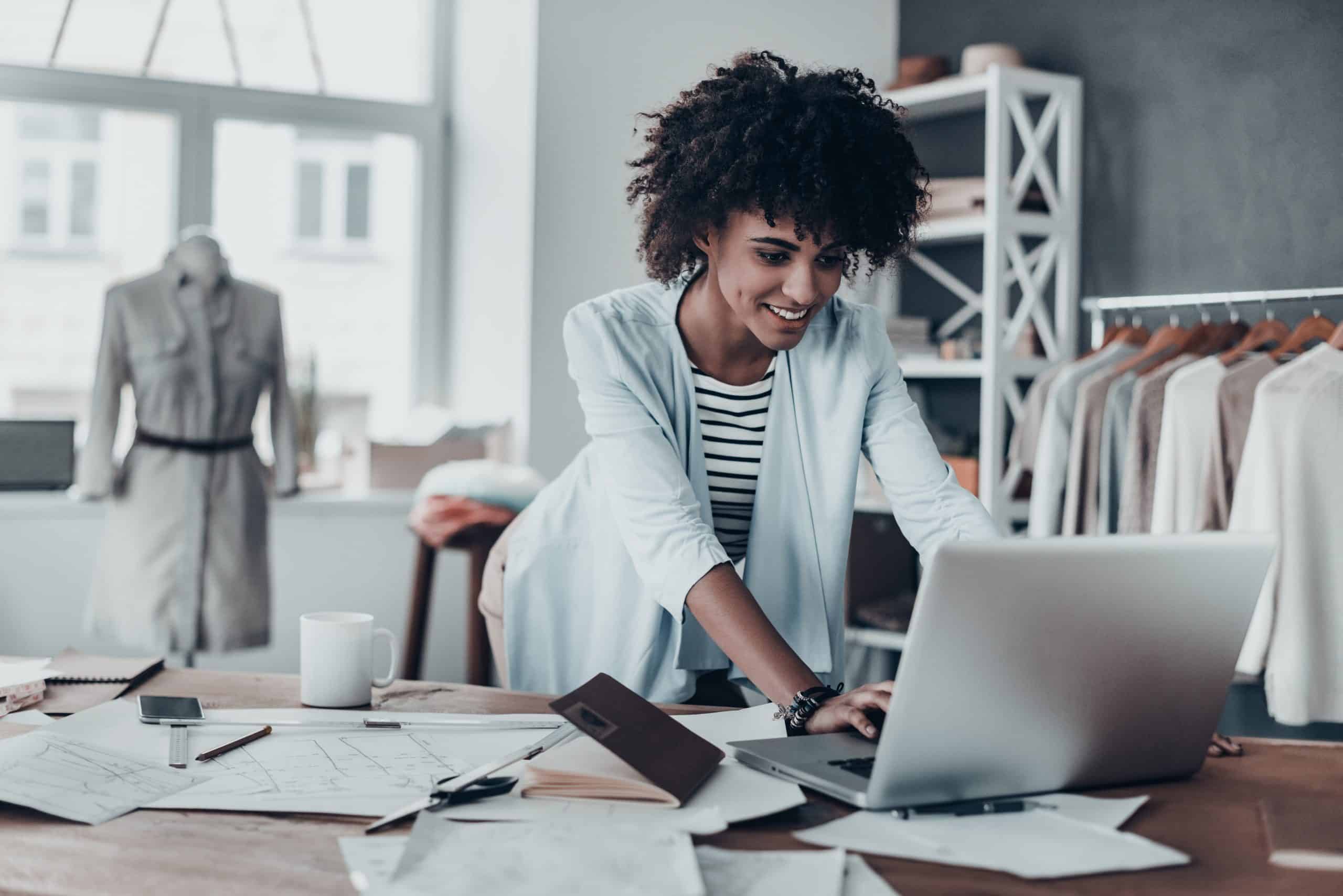 Offices in Huntingdon - photo shows a woman small business owner looking at her laptop at her desk smiling, with papers around her desk and clothes hung on a rail to the side