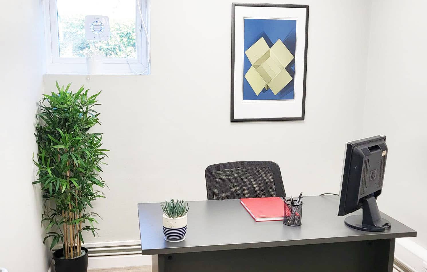 Bicester office space - image shows interior of office with a black desk and plant on the desk with pens and notebook, a green plant in the left hand corner of the room and a painting on the wall behind the desk