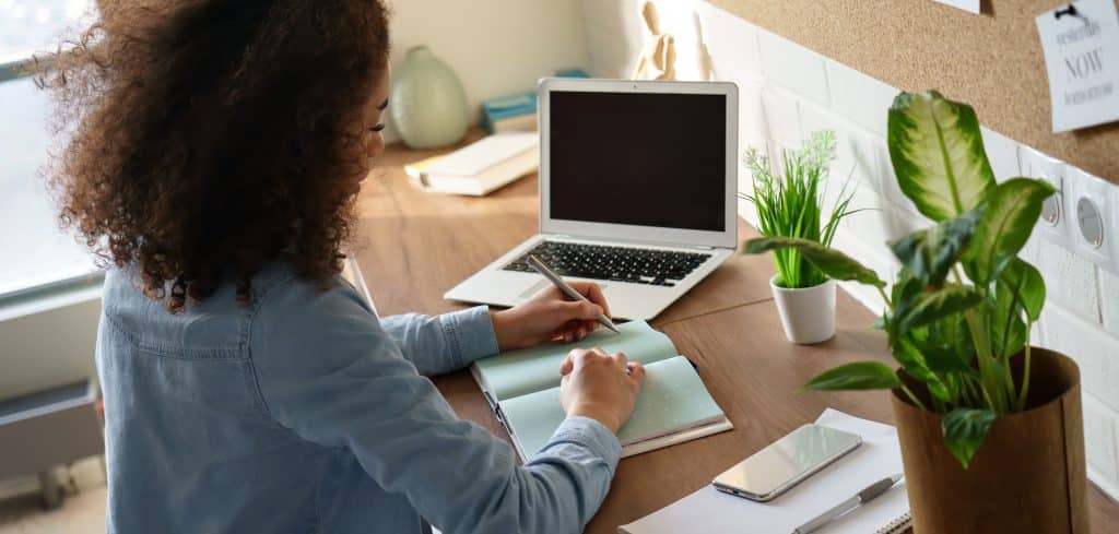 Writing a book: Our Letchworth office space could give you the peace you need. Image shows woman sitting at her desk writing a book with her laptop and green plants on her desk. 