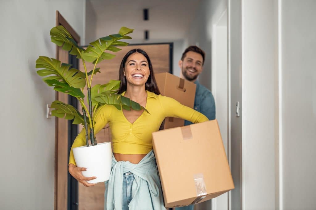 Partner moving in and you need to make room? Our Bicester storage will help. Image shows couple in the hallway carrying boxes and a plant into their home. The woman is wearing a yellow top with jeans and has a jumper tied round her waist. Then man is wearing a jean shirt. 