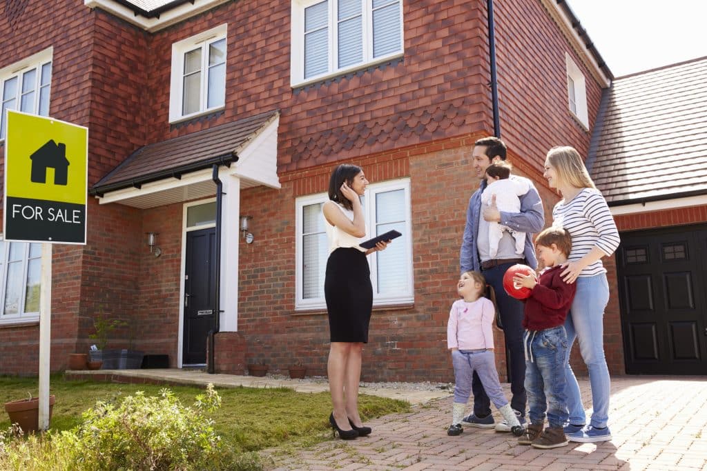 House move storage - image shows a family waiting outside a redbrick house with the estate agent. There is a green 'for sale' sign in the grass