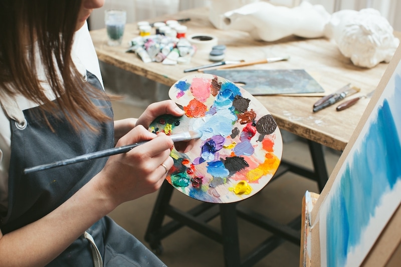 Storage facilities Brentwood. Image shows art student wearing an apron with palette of paint in their hand and a blue painted canvas in front of them 