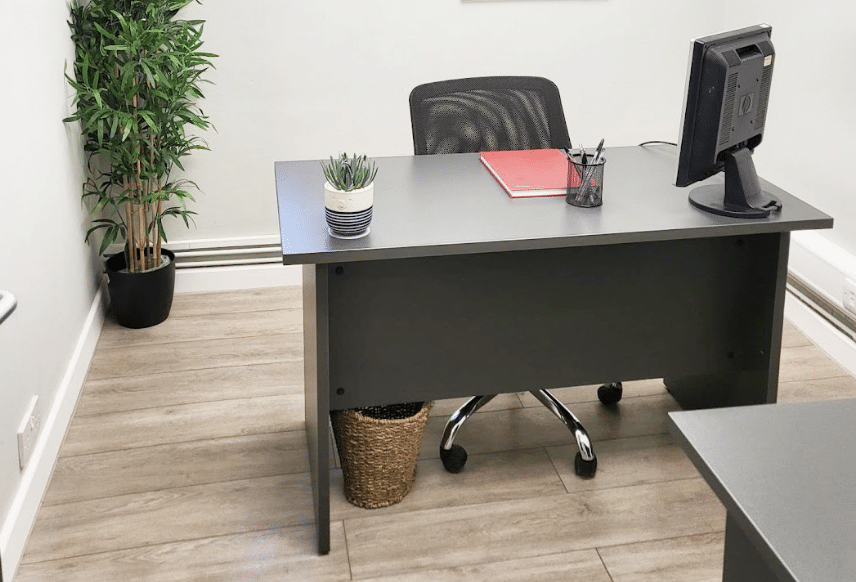 Brentwood office. Image shows a dark grey desk in a room with a plant in the left hand corner and an office chair tucked into the desk. A red notebook is on top of the desk with a pen pot and also small green plant in a pot.