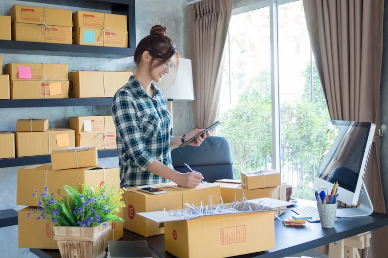 Enfield office space. image shows a woman standing at a desk holding a phone and writing on top of a cardboard box. There are more boxes on the desk with packaging filler and boxes stacked on shelves behind her with a lap to the right. The desk is positioned in front of a large window on the right with brown curtains.