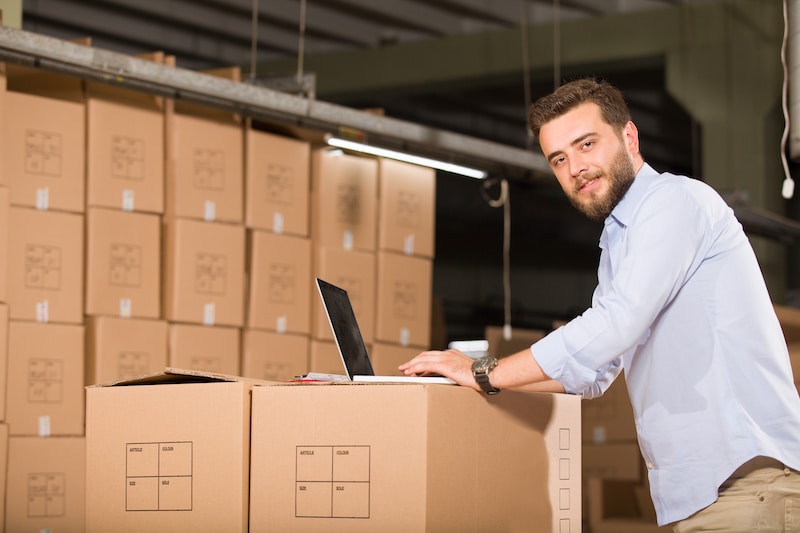 Self Storage in Wandsworth. Image shows a man looking at the camera leaning on his laptop on top of boxes of stock with more boxes stacked up against the wall. 