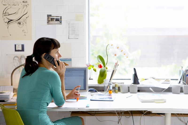 Self storage Brentwood. Image shows a woman at her desk in front of a large window wearing a green dress looking out of the window on the phone with her laptop in front of her.