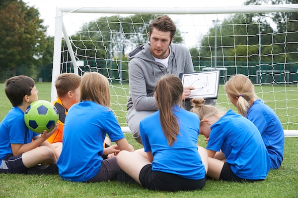 Self storage Watford. Image shows football coach kneeling down in front of goal talking to a group of kids and pointing at his drawing on a whiteboard. 