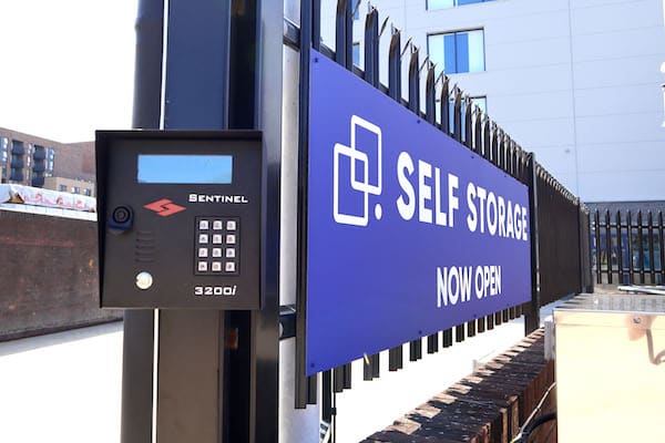 Image shows a blue Cinch Self Storage now open sign on a black fence with a pin code access security box