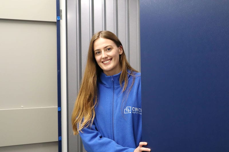 10 benefits of self storage. Image shows a woman in a blue cinch self storage fleece standing at the door of a storage unit smiling. 