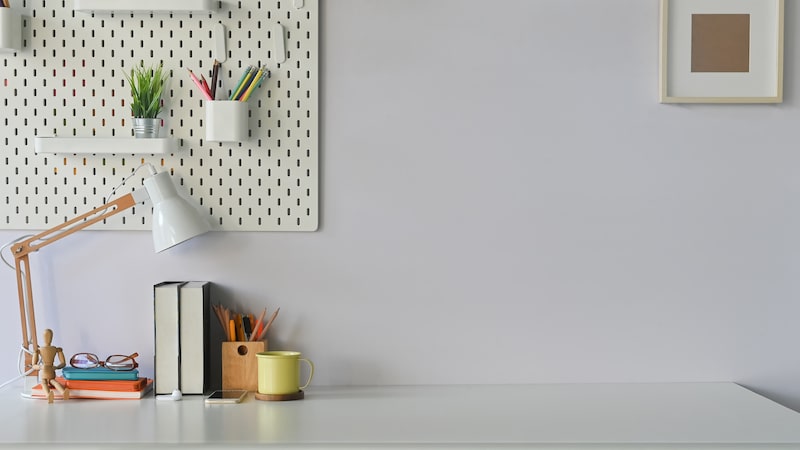 Office to rent in Brentwood. Image shows a desk in an office, with a light grey wall with a pin board with pen pot, white lamp and books with a yellow pencil pot on the desk. 