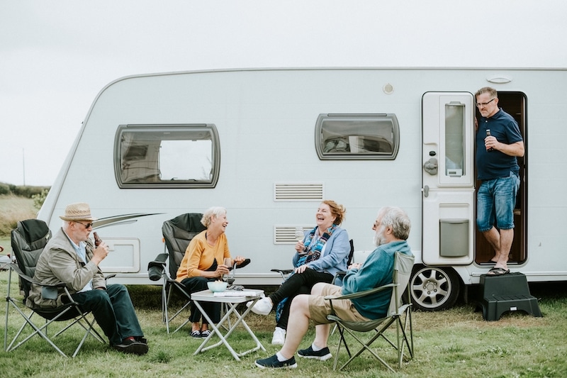 The benefits of using Self Storage for your caravan. Image shows a white caravan in the background with a group of men and women sitting around a camping table laughing. 