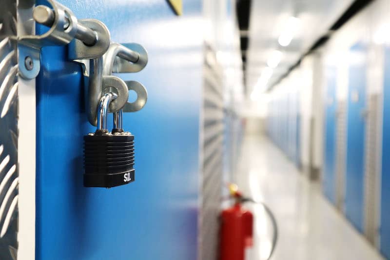 Storage unit in Southend. Image shows a close up of a blue storage unit door with a black padlock, with a corridor of storage units blurred in the background.