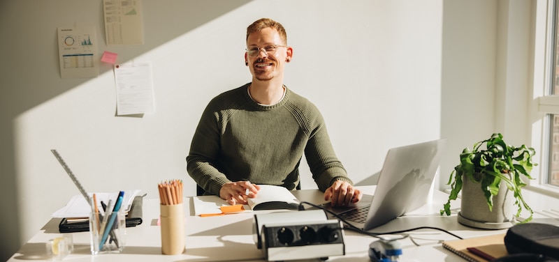 office_to_rent_gillingham.Image shows an entrepreneur wearing a green jumper sitting at his desk with an open laptop and stationery around him.