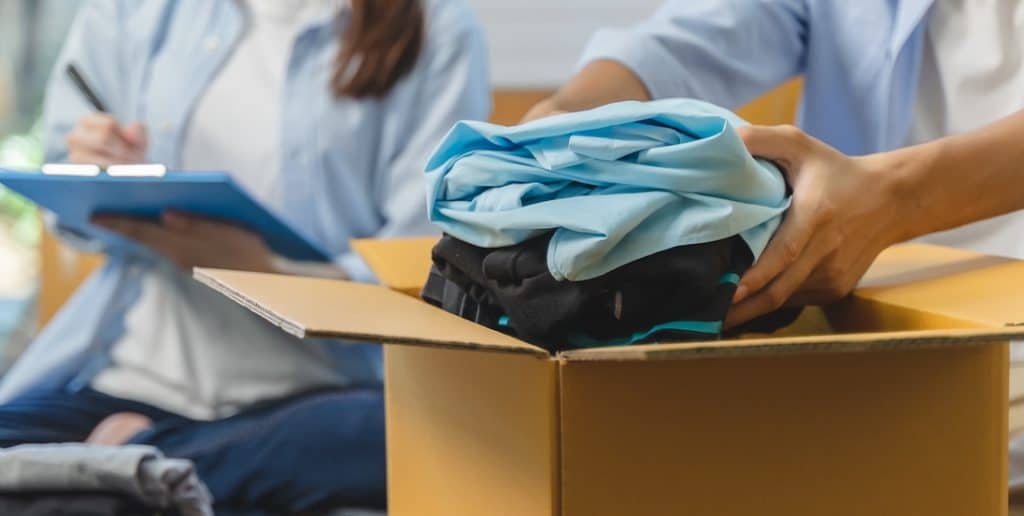 9 Essential tips for storing clothes long-term. Image shows people putting clothes in a cardboard box with someone holding a blue clipboard in the background. 