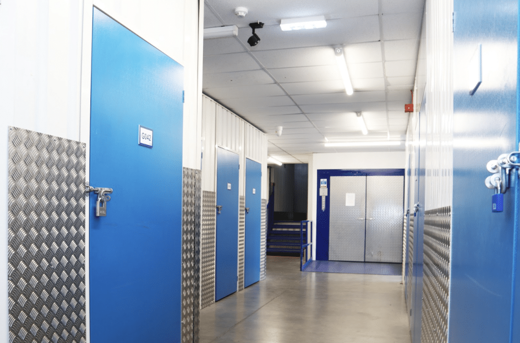 The ultimate guide to renting a storage unit, what you need to know. Image shows blue storage unit doors in a corridor with a lift at the end. 