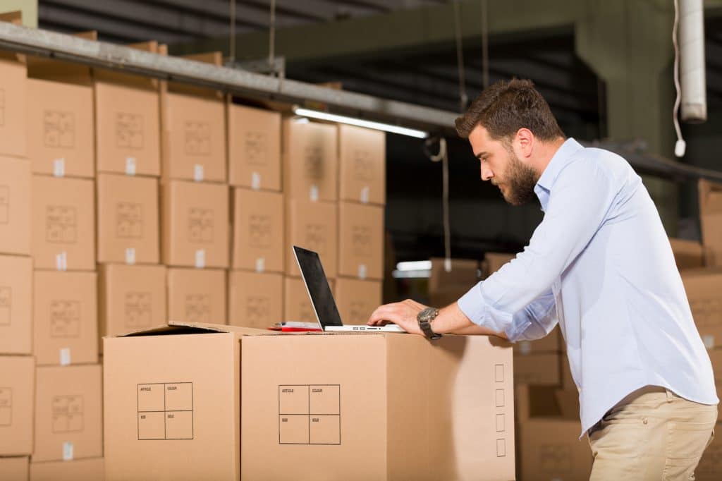 Business storage in Chippenham. Images shows a man in a blue shirt leaning with his laptop on a pile of brown cardboard boxes, with more stacked in front of him. 