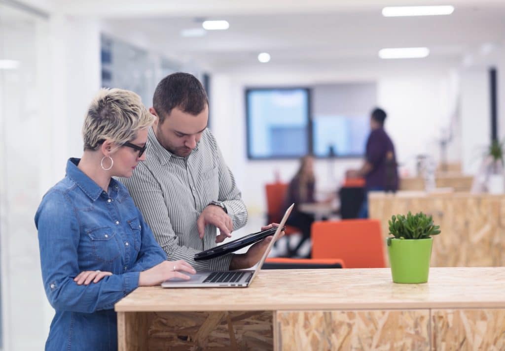 Office to rent in Leighton Buzzard. Image shows a man and a woman pointing on a laptop on a chipboard desk with a white office behind with windows in the back. 