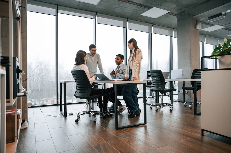 Newmarket office space. Image shows a group of four people sitting and standing round a desk chatting. With large glass windows behind them.