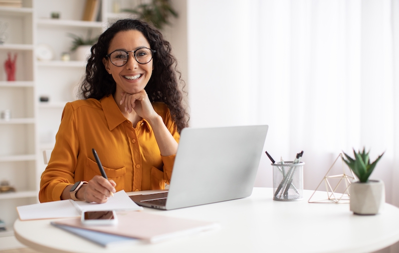 Newmarket office. Image shows a woman in an orange top smiling to camera sitting at her desk with her laptop open.