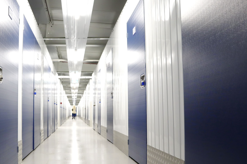 Watford long term storage. Image shows a long corridor of storage unit doors and a woman at the end walking away from the camera. 